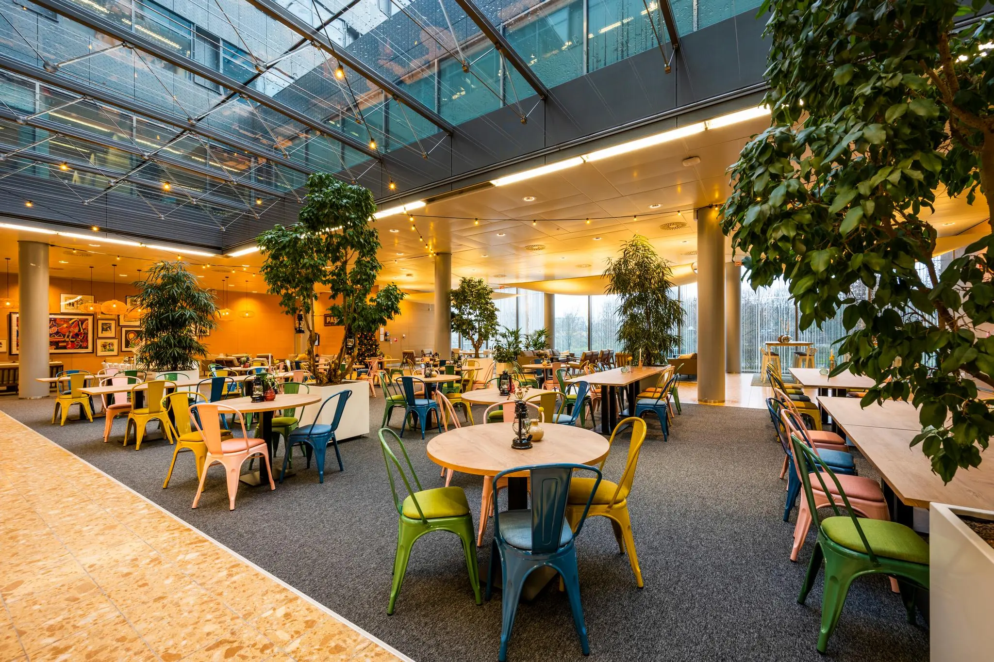 Moderne, kleurrijke bedrijfskantine aan de Burgemeester Rijnderslaan met veel planten en een glazen dak.